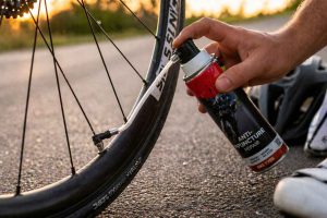 Un cycliste utilise une bombe anti-crevaison sur la valve d'un vélo posé sur une route. Solution de dépannage rapide pour repartir en selle après une crevaison légère, sans démonter le pneu.