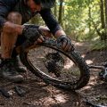 Un cycliste VTT accroupi sur un sentier forestier répare son pneu à plat. Lumière naturelle tamisée à travers les arbres, mains sales, démonte-pneus posés au sol. Guide pratique : que faire en cas de crevaison en pleine sortie vélo.